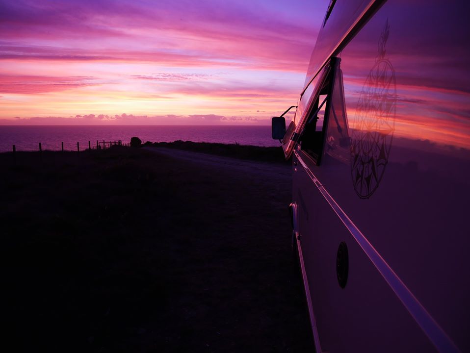 Flanc d'un Volkswagen LT blanc à toit surélevé garé face à l'océan, sous un coucher de soleil rose et violet