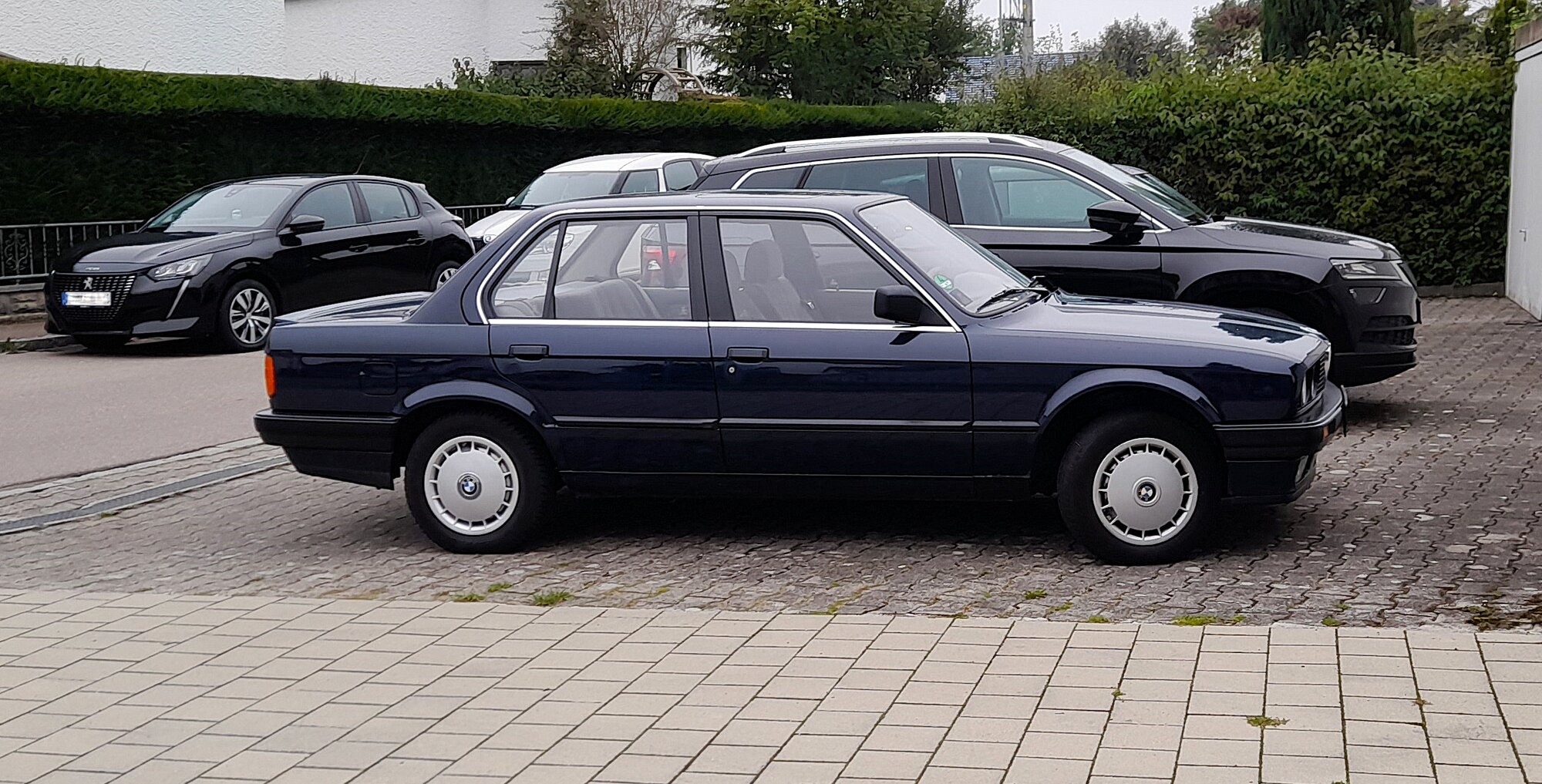 BMW Série 3 E30 phase 2 berline quatre portes bleu nuit vue de profil, garée sur un parking pavé, avec ses enjoliveurs de roue d'origine