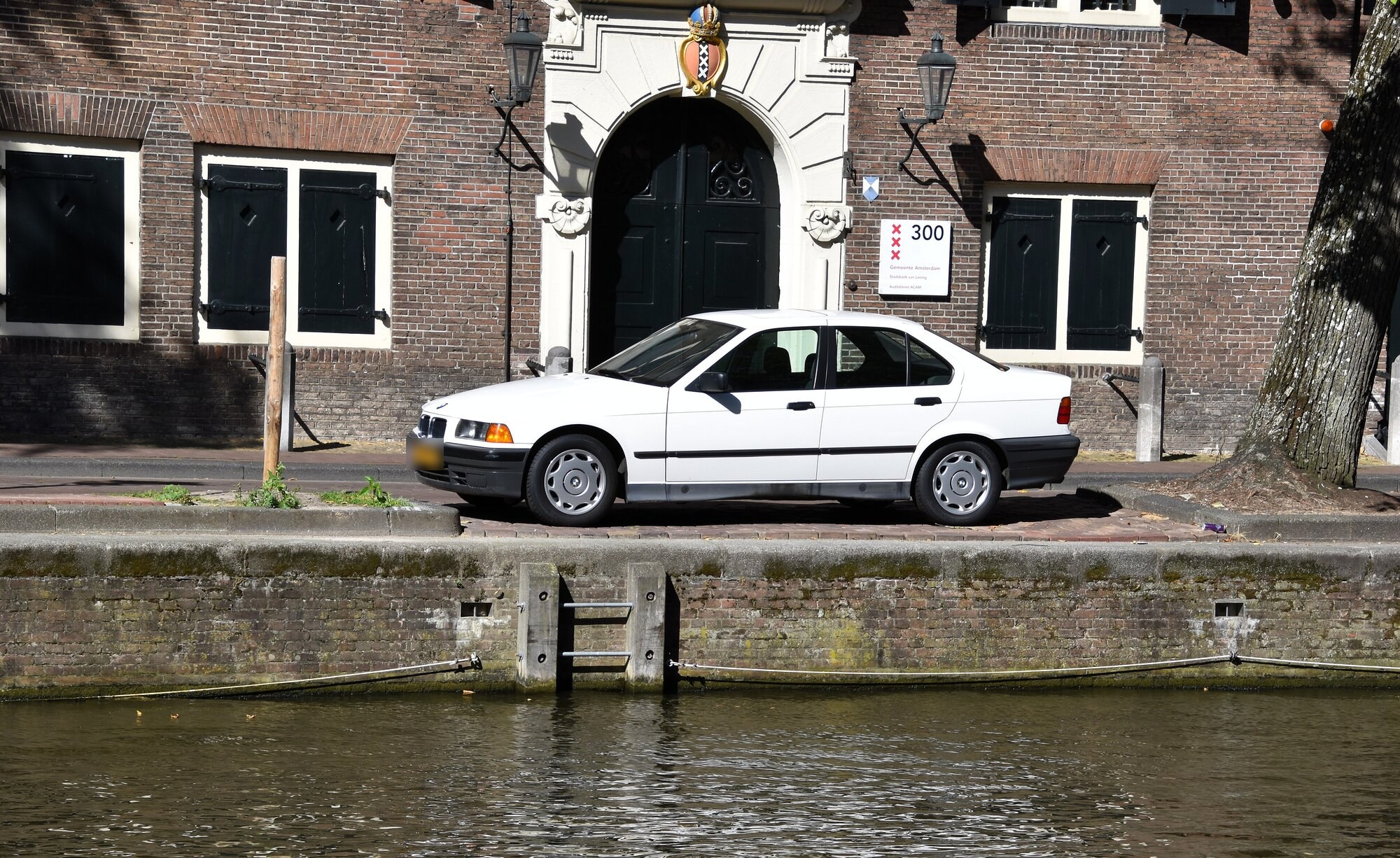 BMW 316i E36 berline blanche de 1993 vue de profil, garée au bord d'un canal devant une façade en briques