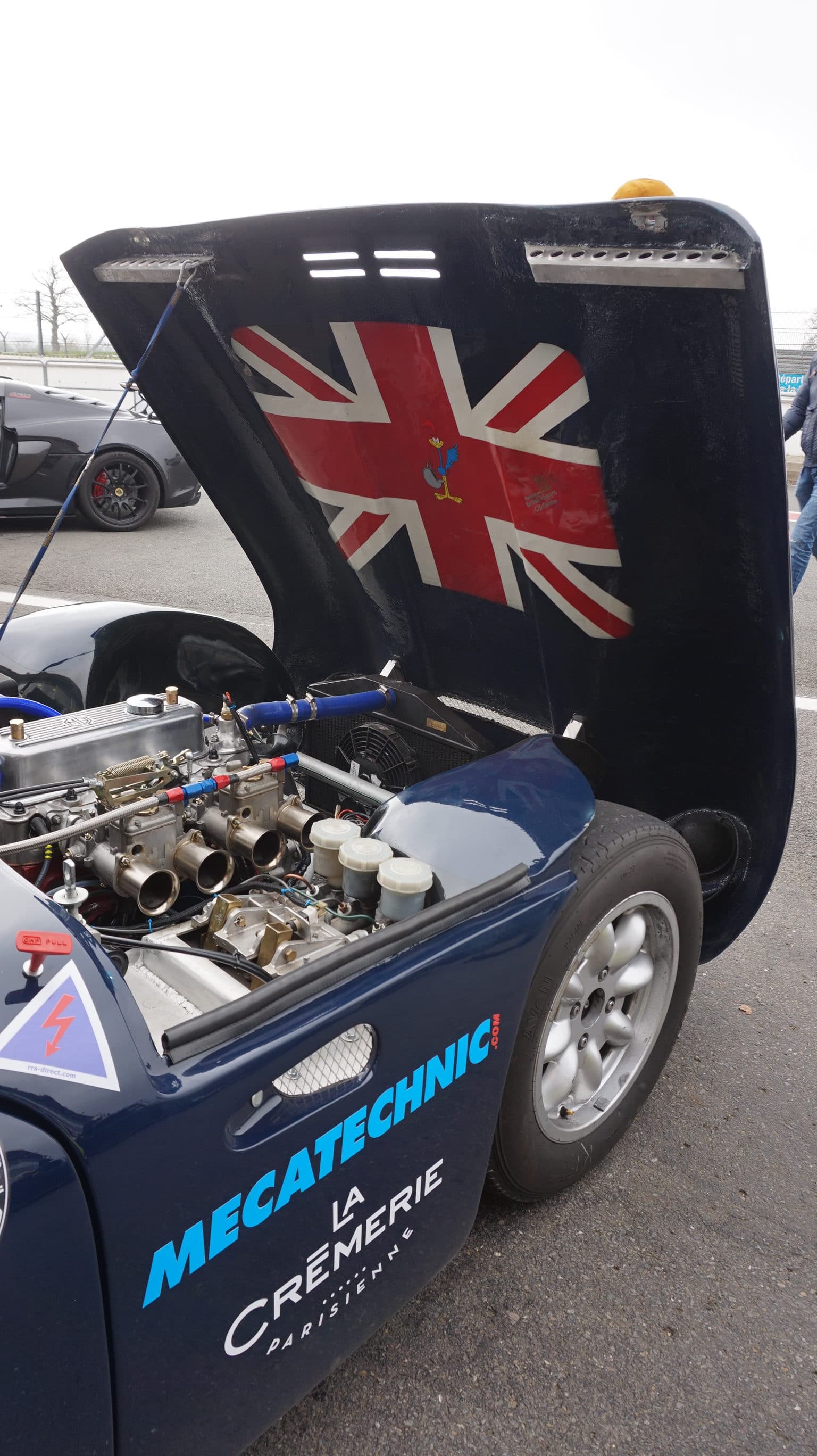 Voiture de course historique bleue capot ouvert sur ses carburateurs, Union Jack peint sous le capot et logo Mecatechnic sur la portière