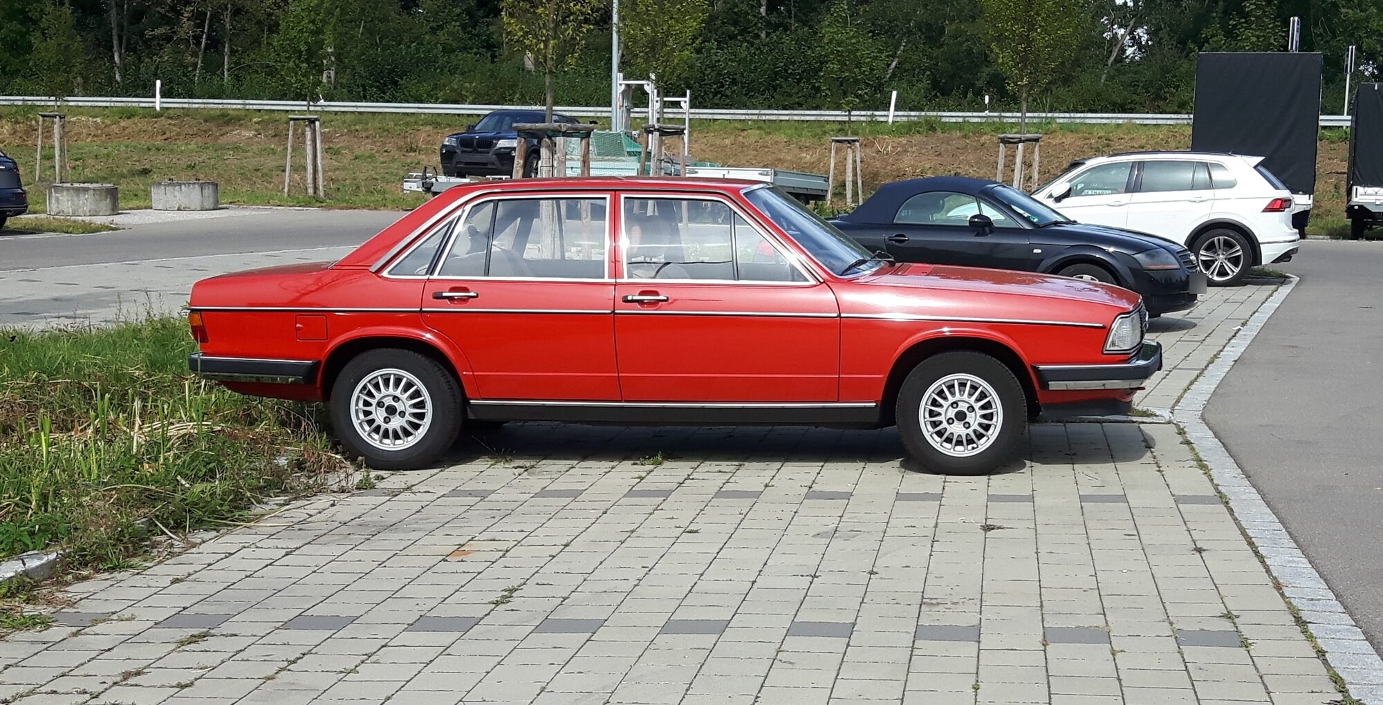 Audi 100 C2 berline rouge vue de profil, garée sur un parking pavé, avec des jantes alliage à rayons