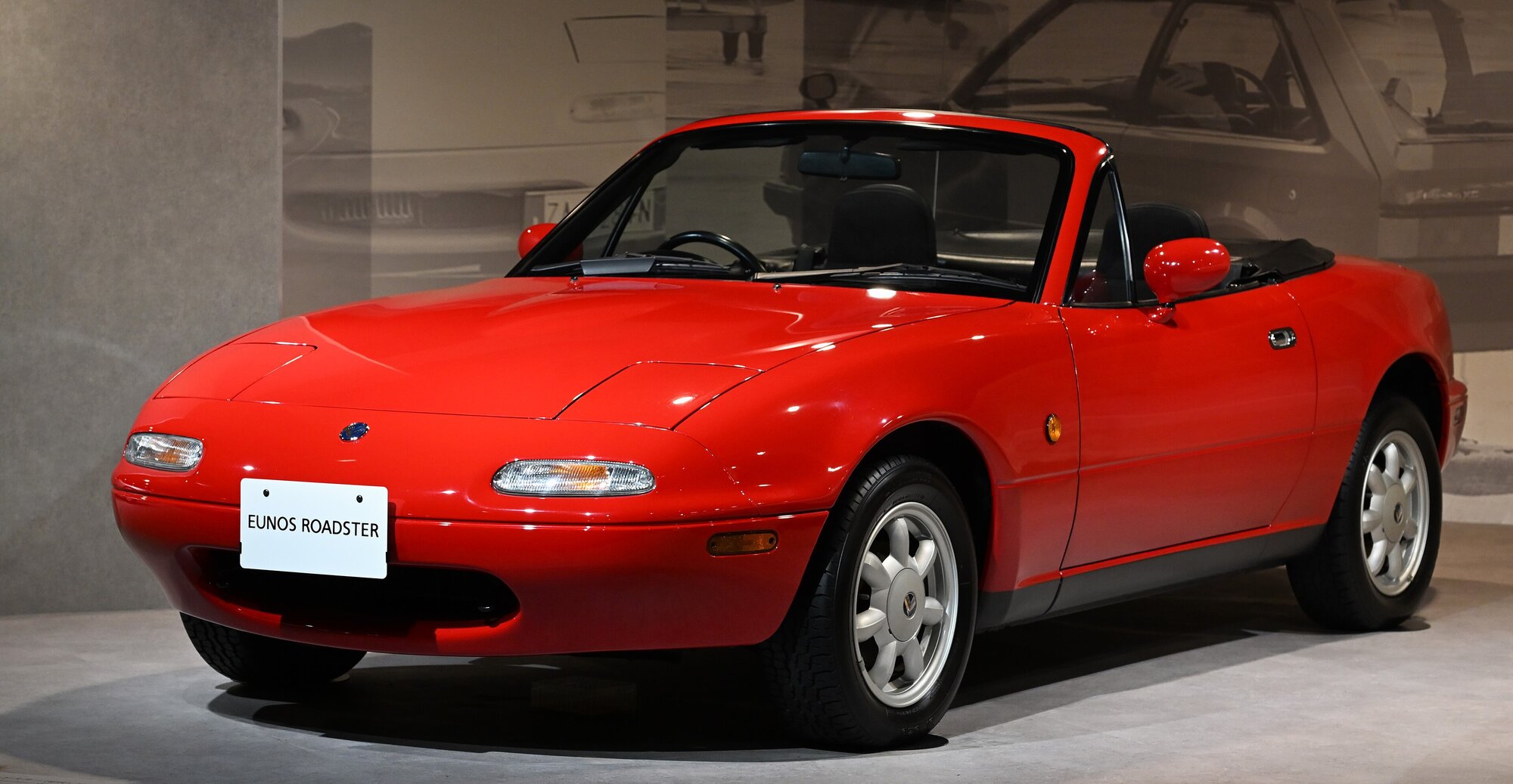 Eunos Roadster rouge de 1989, la MX-5 NA du marché japonais, capote baissée et phares escamotables rentrés, exposée en salle